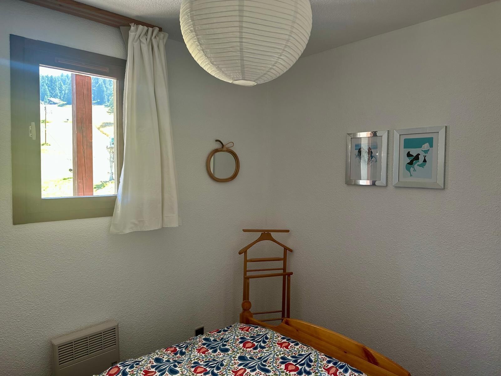 Une chambre à coucher avec un lit, une fenêtre avec des rideaux, un miroir fixé au mur et deux tableaux encadrés.