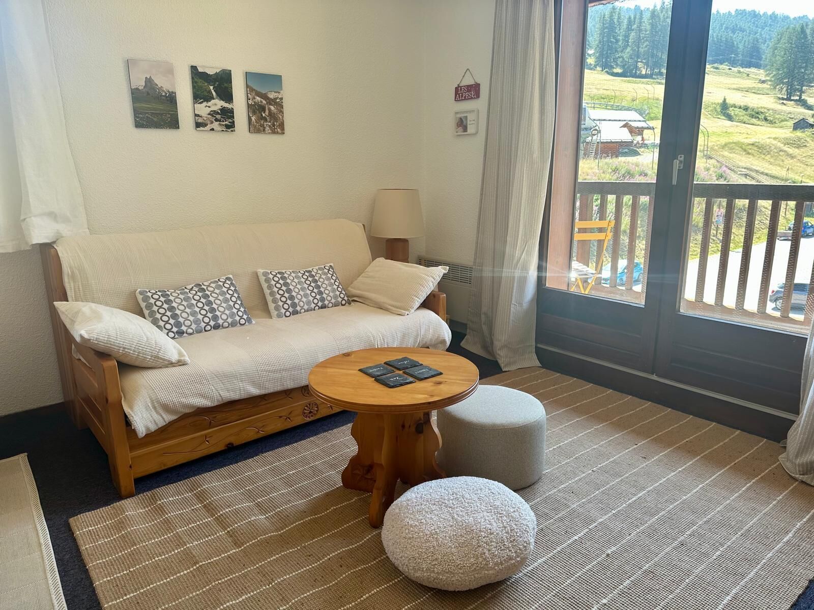 A cozy room with a sofa, a round wooden table, and a view of a balcony with outdoor scenery.