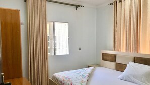 Quarto com cama, mesa de cabeceira de madeira, janela com cortinas e uma porta.