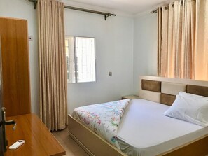 Un dormitorio con cama, una mesita de noche de madera, una ventana con cortinas y una puerta.