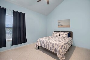A bedroom with a bed, a window with curtains, a ceiling fan, and a painting on the wall. - Seagate Escape condo w. Beach Access (St Augustine)