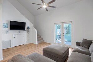 A living room with a flat-screen TV, a ceiling fan, a staircase, and a sliding glass door leading to an outdoor area. - Seagate Escape condo w. Beach Access (St Augustine)