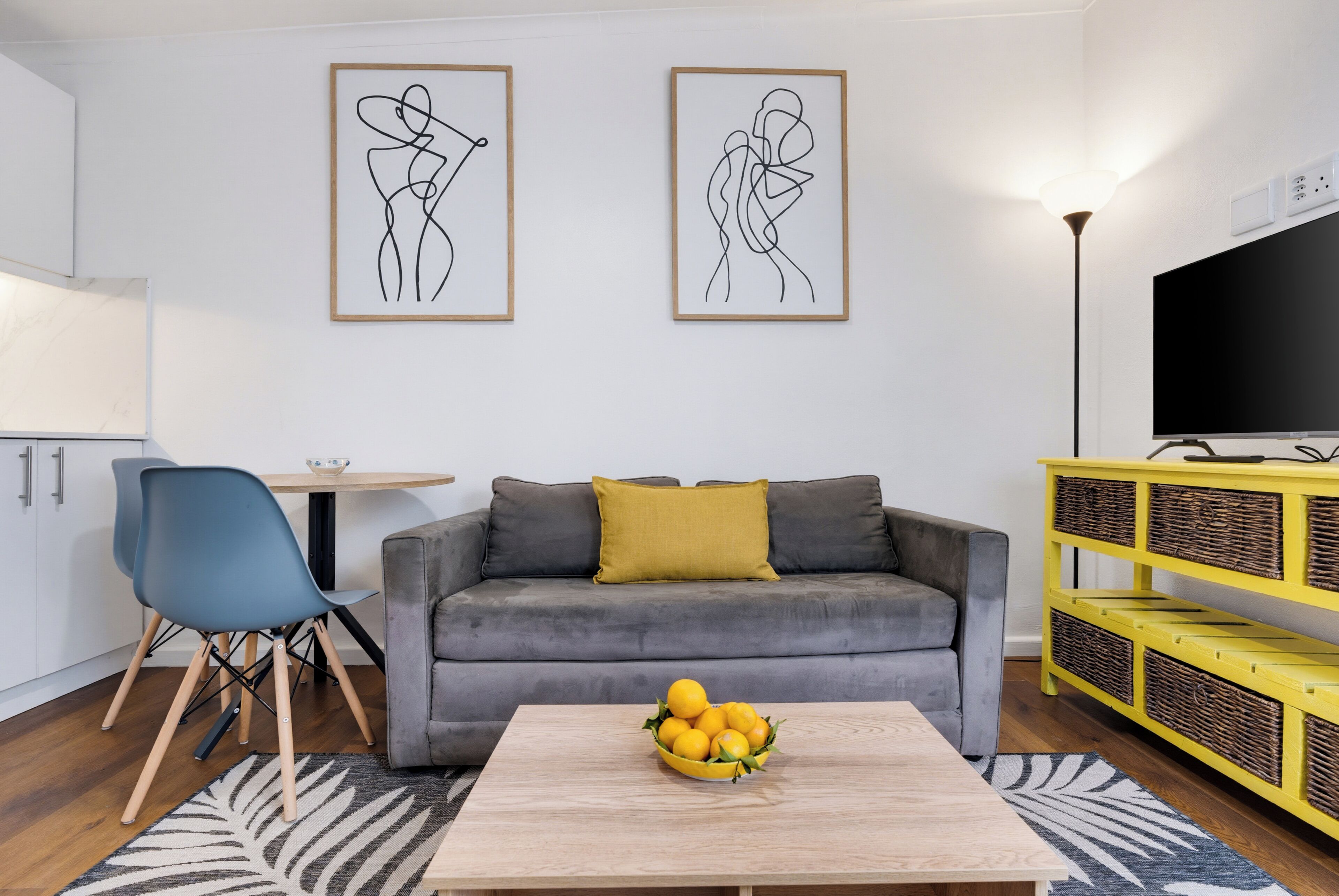 Een woonkamer met een grijze bank, een blauwe fauteuil, een gele consoles en een televisie op een standaard.