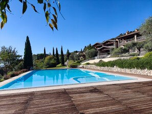 A clear blue swimming pool with a wooden deck in the foreground, surrounded by well-maintained greenery and a hillside with a building in the background. - DELUXE BOGLIACO GOLF APARTMENT MIT 180° - SEEBLICK (Lombardia)