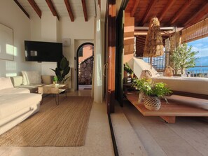 Una sala de estar moderna con sofá, mesa de centro y televisor. Hay un balcón con plantas y vistas al exterior.