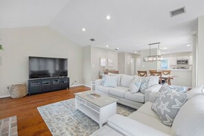 A modern living room with a flat-screen TV, a white sofa set, a coffee table, and a kitchen in the background. - Greenside Getaway - 2BR condo w/ pool access & golf views (Ponte Vedra Beach)