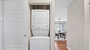 A laundry room with a front-loading washer and dryer, a door leading to a dining area, and a window with blinds. - Greenside Getaway - 2BR condo w/ pool access & golf views (Ponte Vedra Beach)