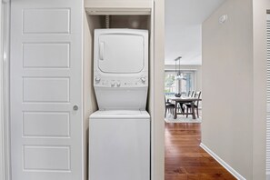 A laundry room with a front-loading washer and dryer, a door leading to a dining area, and a window with blinds. - Greenside Getaway - 2BR condo w/ pool access & golf views (Ponte Vedra Beach)