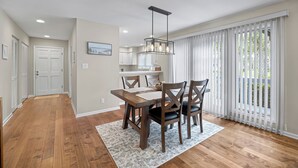 A modern kitchen with white cabinets, a dining area with a wooden table and chairs, and a large window with white blinds. - Greenside Getaway - 2BR condo w/ pool access & golf views (Ponte Vedra Beach)