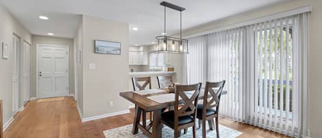 A modern kitchen with white cabinets, a dining area with a wooden table and chairs, and a large window with white blinds.