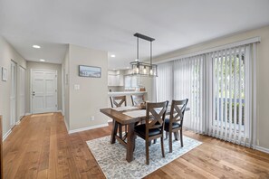 A modern kitchen with white cabinets, a dining area with a wooden table and chairs, and a large window with white blinds. - Greenside Getaway - 2BR condo w/ pool access & golf views (Ponte Vedra Beach)