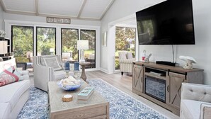 A spacious living room with a large TV, comfortable seating, a coffee table with decorative items, and a view of the outdoors through large windows. - The Sawgrass Sanctuary (PONTE VEDRA BEACH)