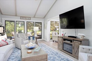 Ein geräumiges Wohnzimmer mit großem Fernseher, bequemen Sitzgelegenheiten, einem Couchtisch mit Dekoration und Blick ins Freie durch große Fenster.