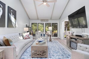 Un salon spacieux avec un ventilateur de plafond blanc, un grand téléviseur à écran plat, un canapé blanc, une table basse agrémentée d’objets décoratifs et un tapis à motifs.