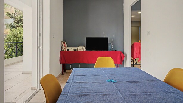 Um quarto com uma mesa azul, cadeiras amarelas, uma mesa coberta por uma toalha vermelha e equipamentos de áudio, e uma parede cinza.