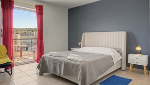 Quarto com cama, mesa de cabeceira com abajur, cadeira, varanda com vista e um tapete azul.
