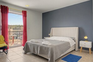 Ein Schlafzimmer mit Bett, Nachttisch mit Lampe, Sessel und Balkon mit Blick, sowie einem blauen Teppich.