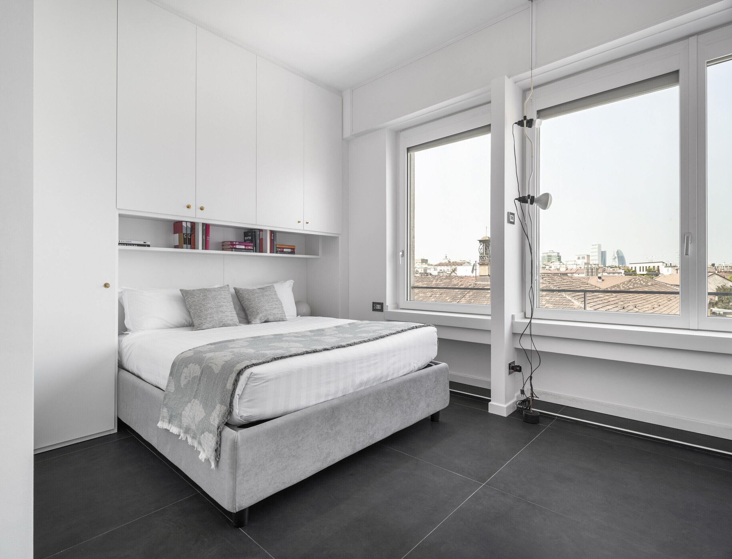 Un dormitorio moderno con cama, estanterías empotradas, un ventanal y vistas a la ciudad.