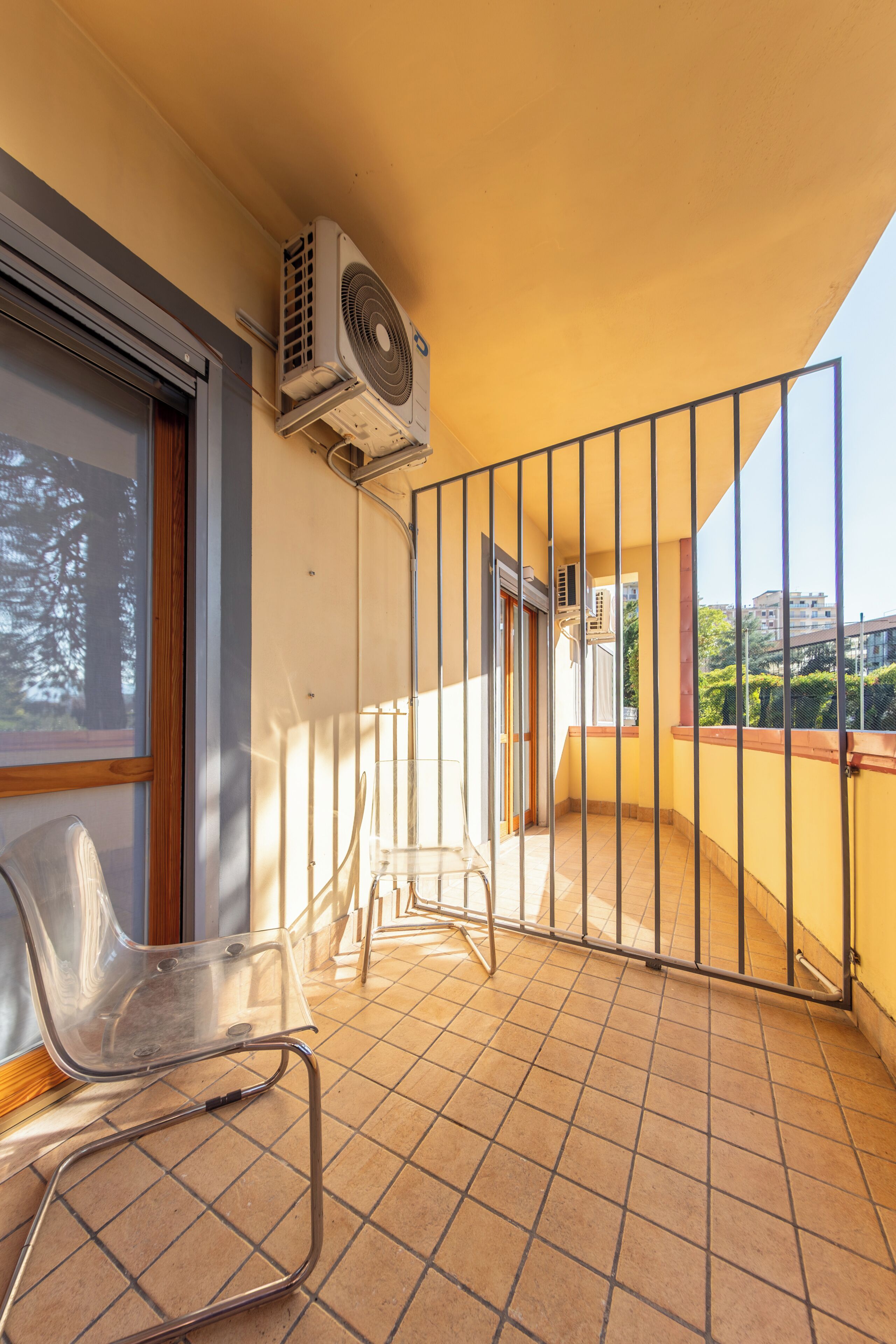 A balcony with a metal gate, a clear glass chair, and an air conditioning unit mounted on the wall.