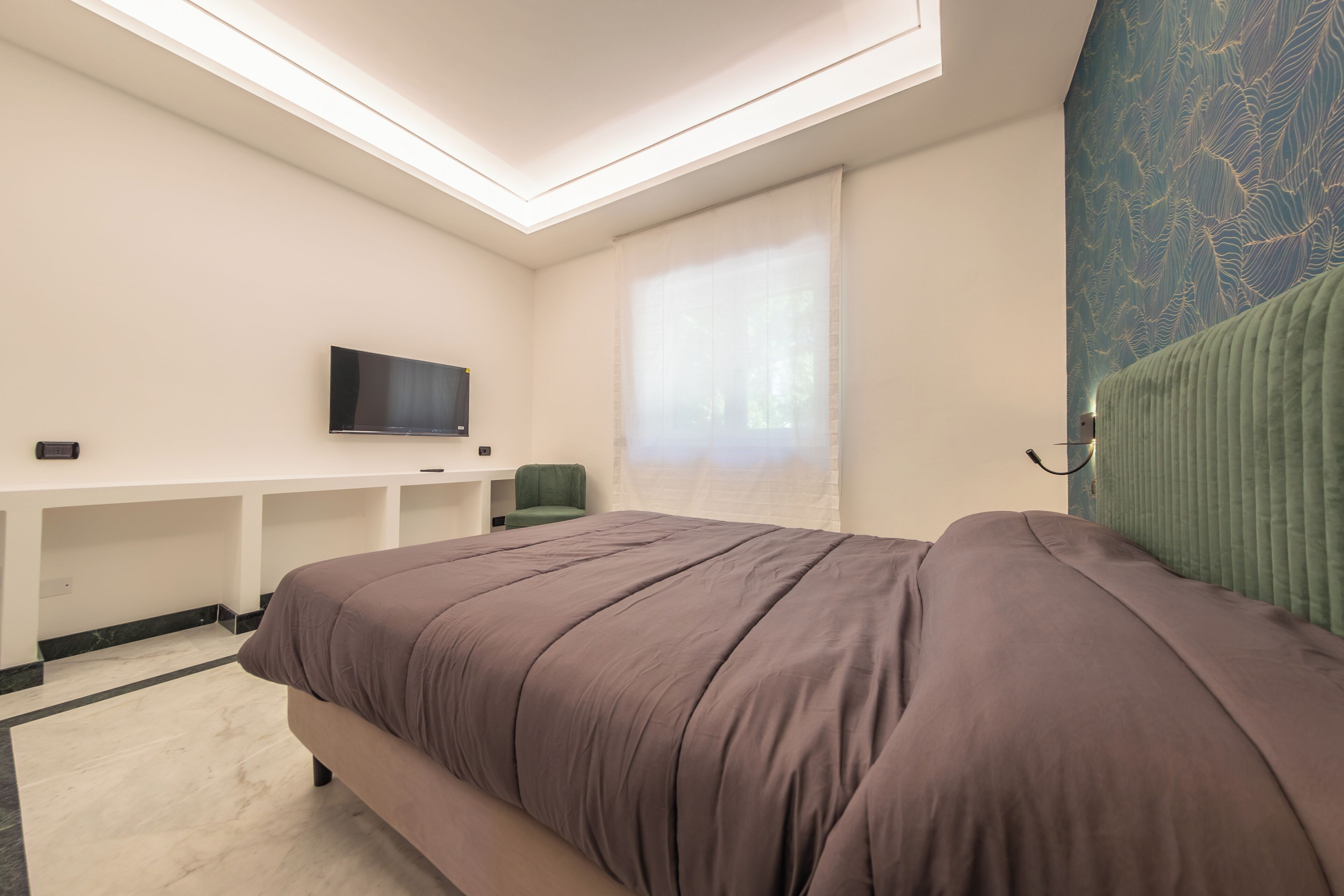 Hotelkamer met een bed, een bureau met een flatscreen televisie, een raam met dunne gordijnen en een behang met een patroon.