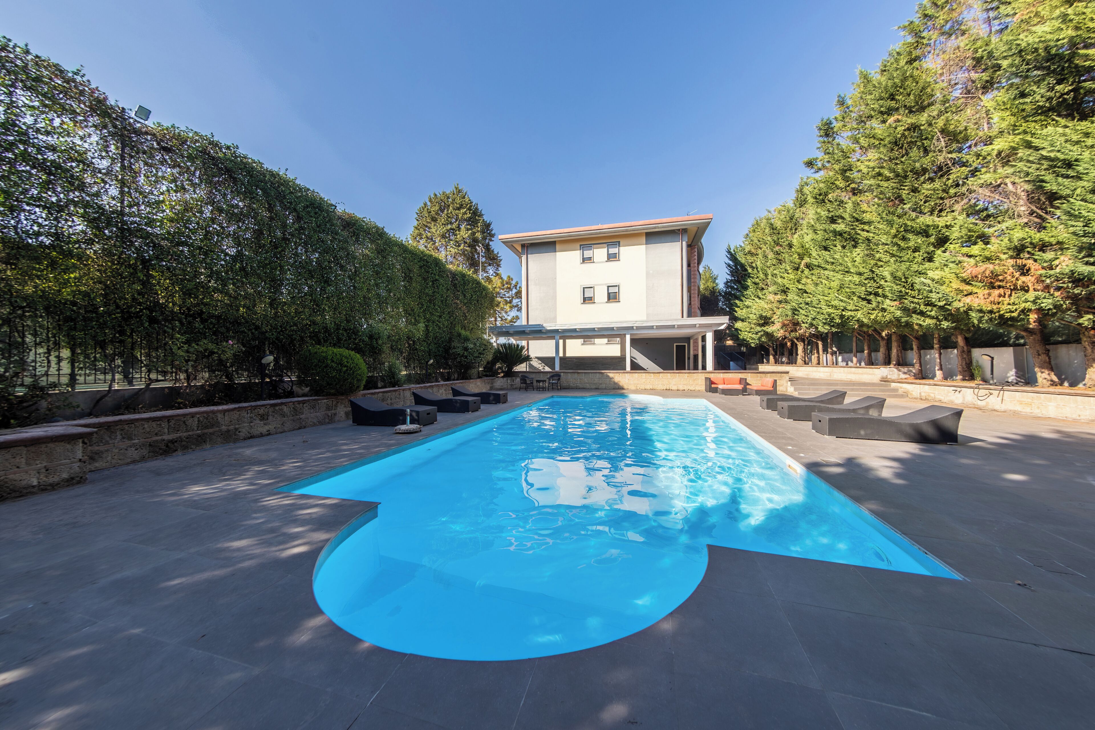 A rectangular swimming pool with a clear blue water surface, surrounded by a paved deck. There are several lounge chairs and a small table on the deck. A building with a flat roof and multiple windows is visible in the background.