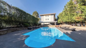 A rectangular swimming pool with a clear blue water surface, surrounded by a paved deck. There are several lounge chairs and a small table on the deck. A building with a flat roof and multiple windows is visible in the background.