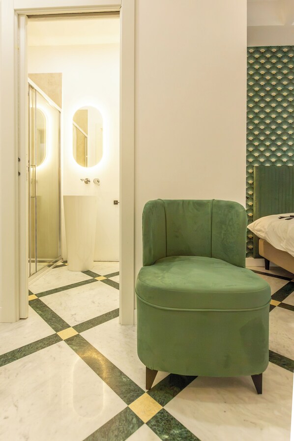A room with a green ottoman, a bed with a white bedspread, a mirror with a round light, and a patterned wall.