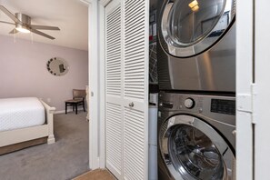 A bedroom with a bed, a chair, a ceiling fan, a wall clock, and a washing machine. - Phoenix Iii 3071 3 Bedroom Condo (Orange Beach)