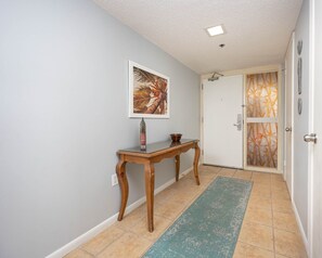 A hallway with a wooden console table, a framed picture, a door with a patterned glass panel, and a green mat on the floor. - Phoenix Iv 4085 3 Bedroom Condo (Orange Beach)