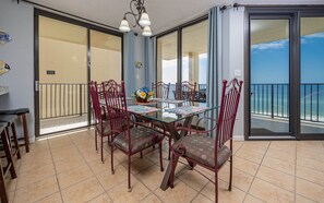 A dining area with a glass table and chairs, a chandelier, and a view of the ocean through large sliding glass doors. - Phoenix Iv 4085 3 Bedroom Condo (Orange Beach)