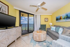 A living room with a flat-screen TV, a round coffee table, a sofa, a ceiling fan, and a balcony with a view of the ocean. - Phoenix Iii 3098 2 Bedroom Condo (Orange Beach)