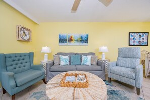 Ein Wohnzimmer mit einem blauen Sofa, einem grauen Sofa und einem gemusterten Sessel. Zwei Beistelltische mit Lampen, ein Couchtisch mit Dekoration und ein Gemälde an der Wand sind ebenfalls vorhanden.