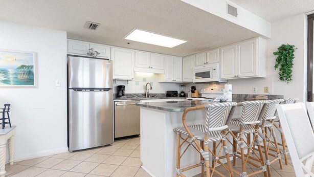 A modern kitchen with white cabinetry, stainless steel appliances, and a bar area with wicker chairs. - Phoenix Iv 4075 3 Bedroom Condo (Orange Beach)