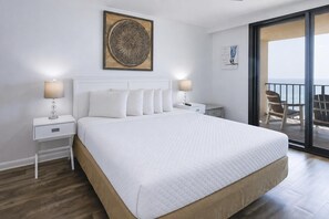 Quarto de hotel com uma cama grande, duas mesas de cabeceira, um criado-mudo, um objeto decorativo na parede e vista para o exterior através de uma porta de correr.