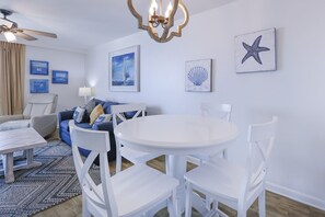 A room with a round white table, white chairs, a blue sofa, a ceiling fan, and nautical-themed wall art. - Phoenix Iii 3068 2 Bedroom Condo (Orange Beach)