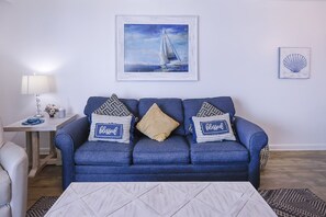 Un salon avec un canapé bleu, deux coussins décoratifs, un tableau encadré représentant un voilier et une petite table sur laquelle se trouvent une lampe et des fleurs.