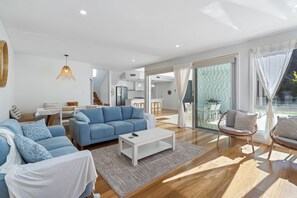 A modern living room with a blue sofa, white armchairs, a coffee table, and a dining area with a table and chairs. - 17 Elluisa Close : 4  BR, 3  BA House in Forster, Sleeps 8 (Forster)
