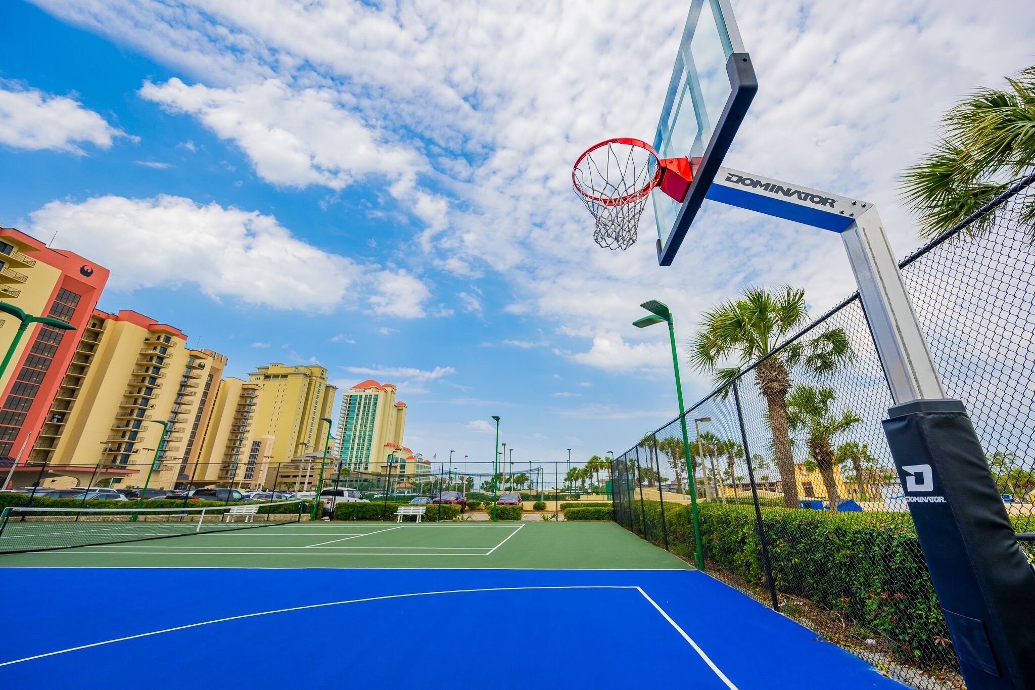 Ein Basketballplatz mit Netz, umgeben von einem Zaun, Palmen und Gebäuden im Hintergrund.