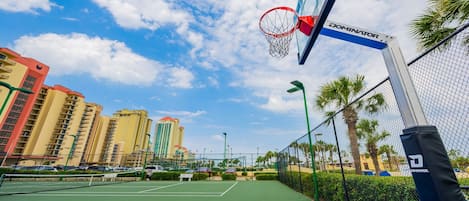 A basketball court with a net, surrounded by a fence, palm trees, and buildings in the background.