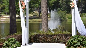 A white gazebo with draped fabric and floral decorations, a pond with a water feature, and surrounding greenery. - 501l Studio (Calabash)
