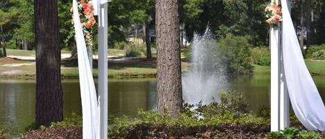 A white gazebo with draped fabric and floral decorations, a pond with a water feature, and surrounding greenery.