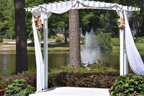 Ein weißes Gazebo mit herabhängenden Stoffbahnen und floraler Dekoration, ein Teich mit Wasserspiel und umgebende Grünflächen.