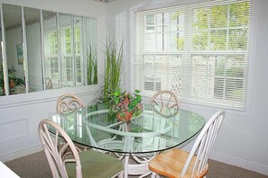 A dining area with a glass table surrounded by white chairs, a potted plant, and a window with blinds. - 501m 1 Bedroom Condo (Calabash)