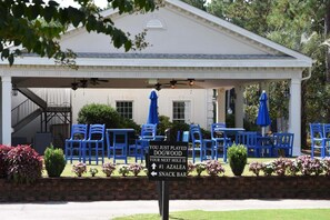 Un espace extérieur abrité avec des chaises bleues et des parasols, un panneau indiquant la direction du bar Azalea, et un bâtiment blanc avec un toit à pignon en arrière-plan.