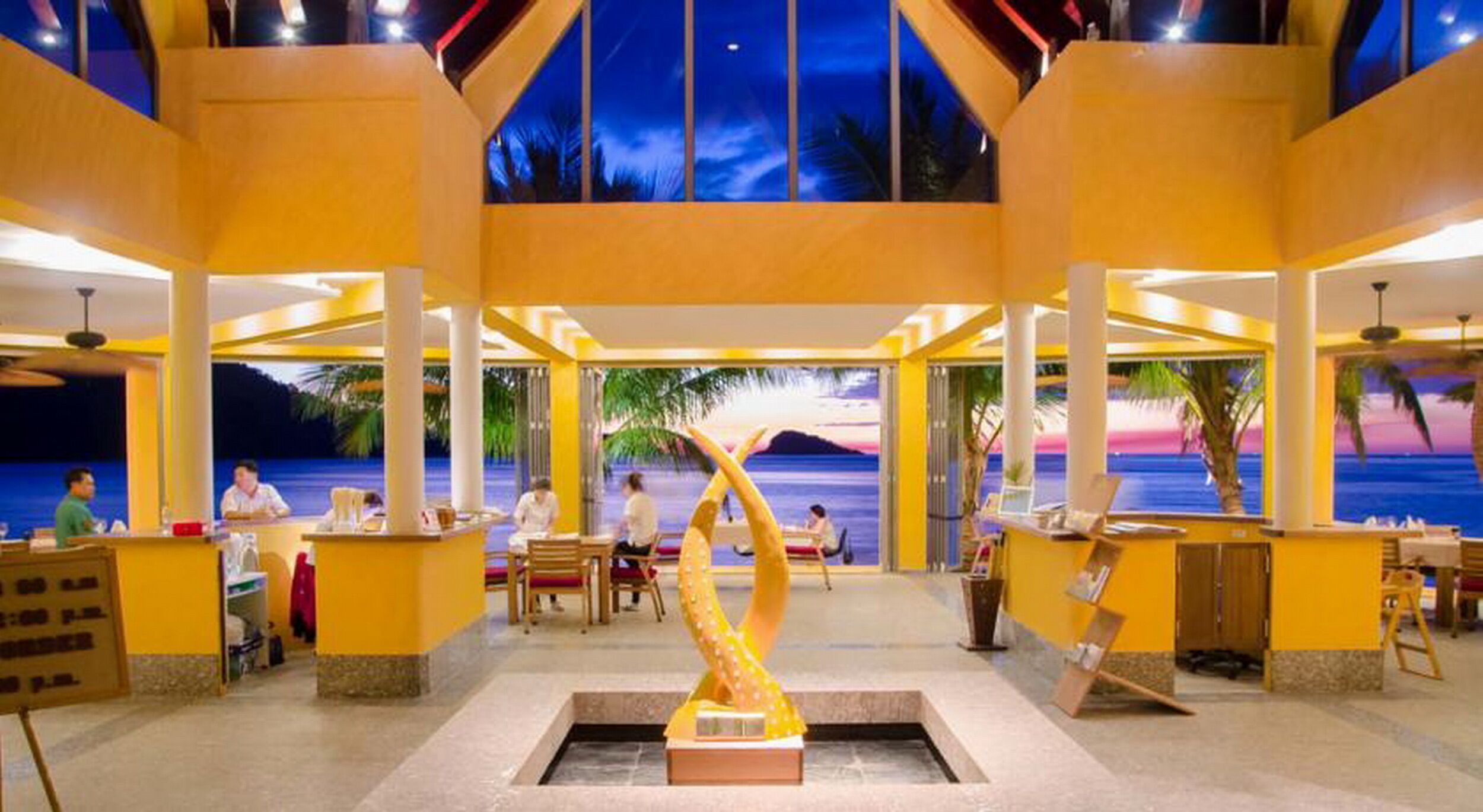 A spacious indoor area with a central sculpture, multiple seating arrangements, and a view of the ocean through large windows.