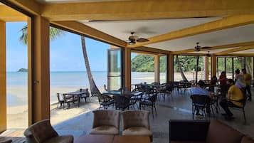 A covered outdoor seating area with tables and chairs, overlooking a beach and ocean.