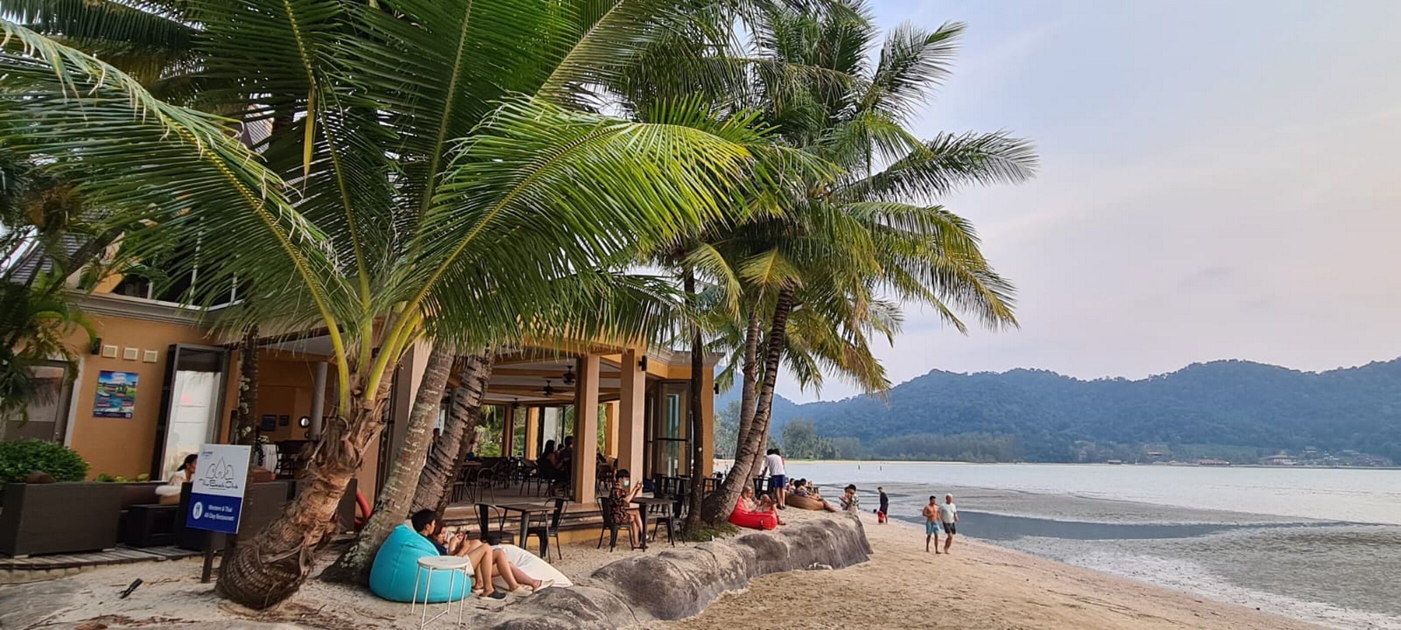 A beachside cafe with outdoor seating, palm trees, and a view of the sea.