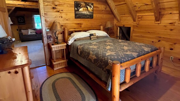 Un dormitorio de cabaña de madera con una cama, una mesita de noche, una lámpara, un ventilador de techo y un cuadro en la pared.
