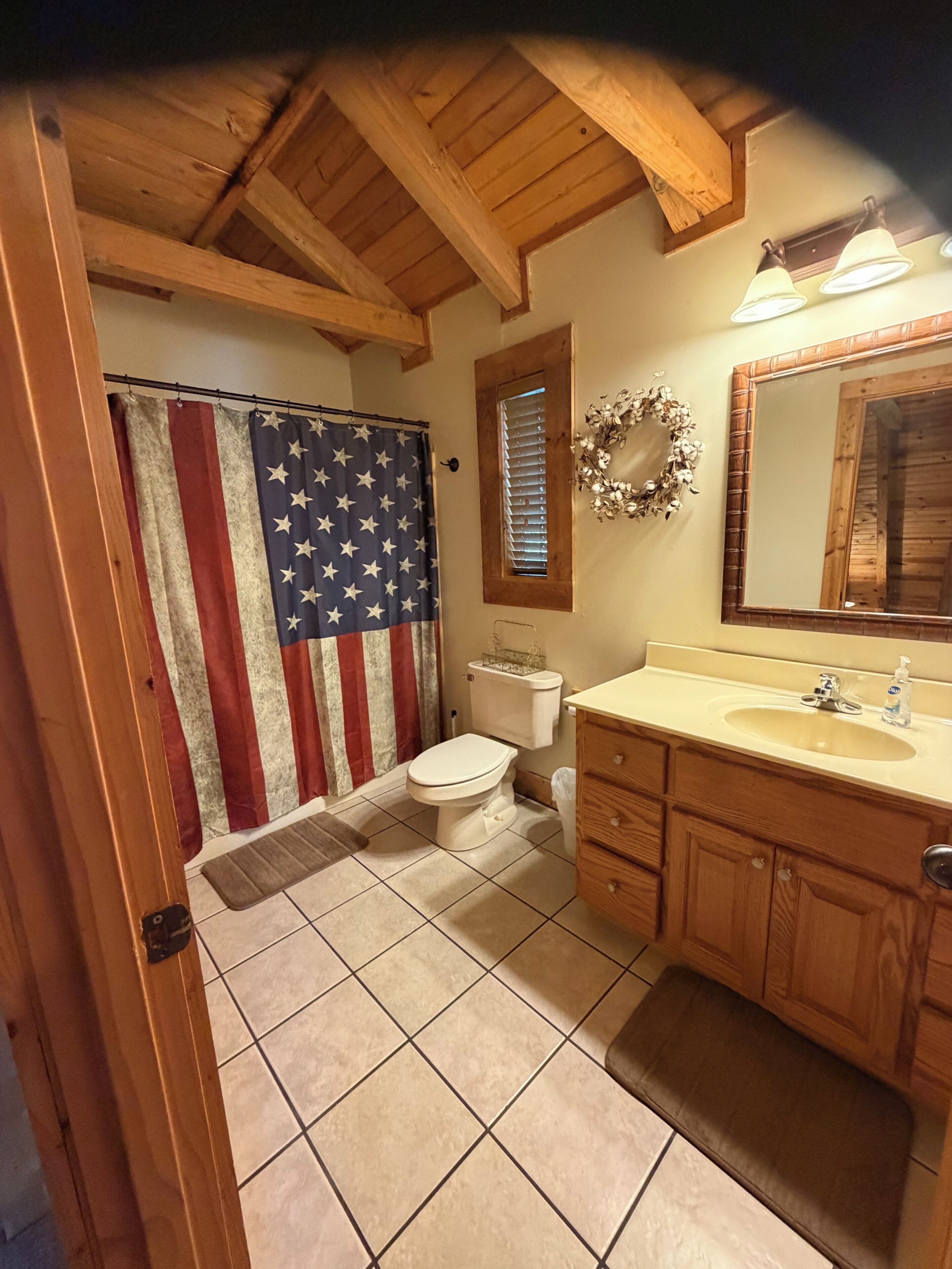 Un baño con techo de madera, un inodoro, un lavamanos con espejo y una cortina con la bandera estadounidense.