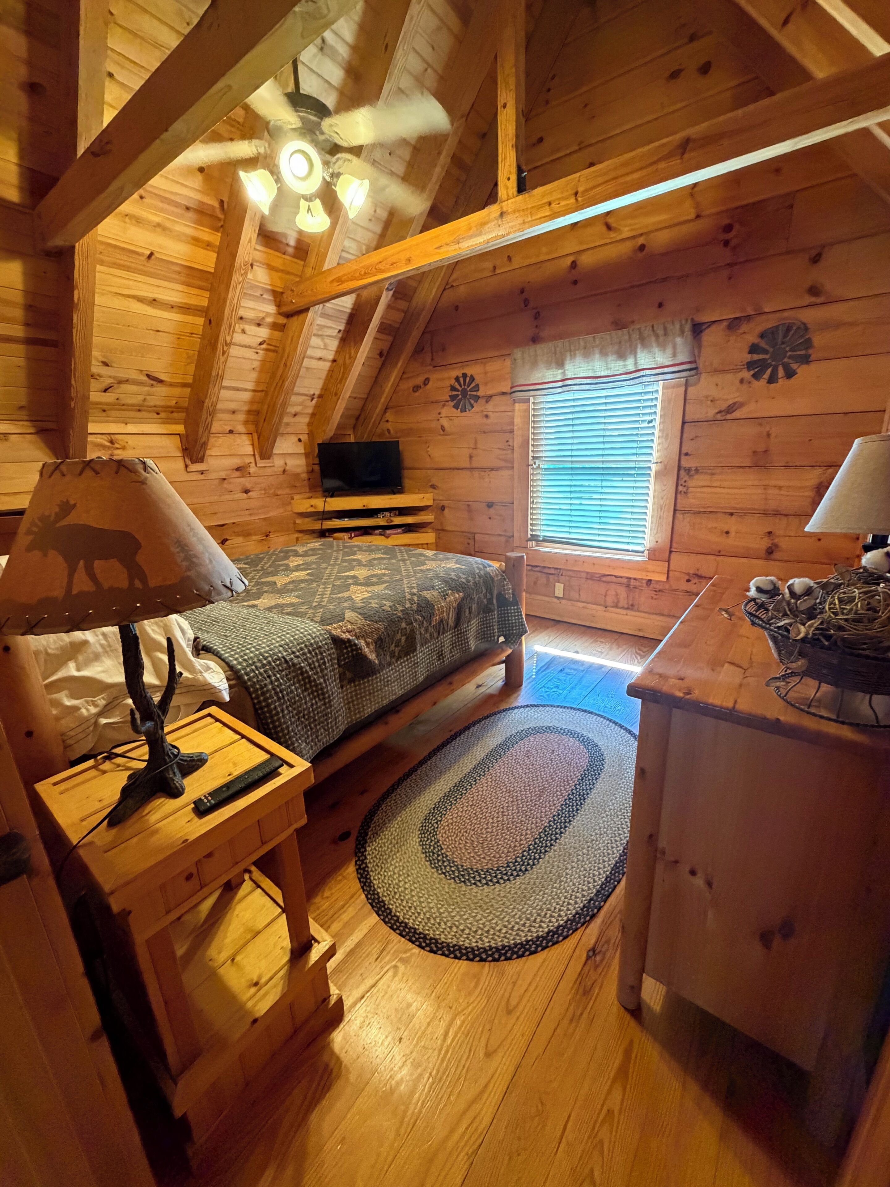 Une chambre confortable dans un chalet, avec un lit, un bureau, une lampe, une télévision et une fenêtre équipée de stores.
